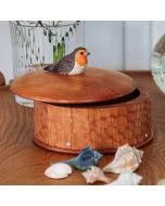 Ronde houten bewaardoos met decoratief roodborstje op het deksel, geplaatst op een houten tafel met schelpen en een glazen vaas op de achtergrond.