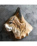 Houten lynxkop als magneet op een donkere metalen wand, met lange zwarte oorpunten, bruingevlekte kop en lichte snuit met donkere stippen.