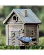 Boomklever met blauwgrijze rug en zwarte oogstreep hangt bij de voederopening van een houten vogelhuis in Scandinavische stijl, met geribbeld dak en decoratieve deur met visgraatpatroon.