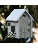 Koolmees eet vogelvoer bij de Multiholk Modern Nordic van Wildlife Garden, een grijs houten nestkast en voederhuis met lichtgrijs zadeldak op een tuinpaal.