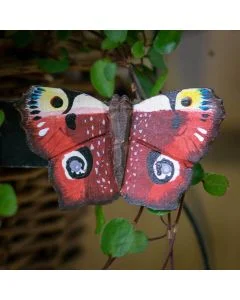 Decoratieve houten vlinder in de vorm van een dagpauwoog, met vier kenmerkende oogtekeningen en donker omrande roodbruine vleugels tussen groene takjes.