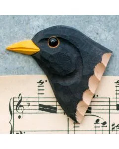Decoratieve houten vogelmagneet in de vorm van een merel, met helder gele snavel en zwarte veren, liggend op een muziekblad met noten.