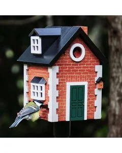 Houten vogelhuis in Engelse cottage-stijl met een koolmees op de zitstok, rode baksteenprint, donkergroene deur en witte ramen tegen een donkere tuinachtergrond.