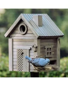 Boomklever met blauwgrijze rug en zwarte oogstreep hangt bij de voederopening van een houten vogelhuis in Scandinavische stijl, met geribbeld dak en decoratieve deur met visgraatpatroon.