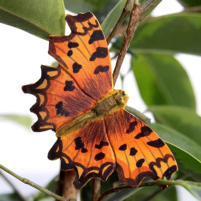 Decoratieve houten vlinder in de vorm van een gehakkelde aurelia, met feloranje vleugels, onregelmatige zwarte vlekken en diep ingesneden vleugelranden tussen planten.