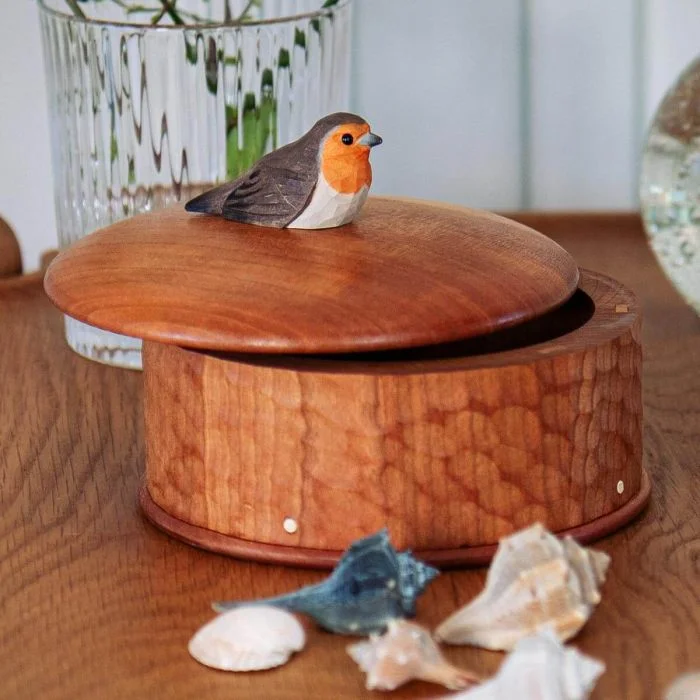 Ronde houten bewaardoos met decoratief roodborstje op het deksel, geplaatst op een houten tafel met schelpen en een glazen vaas op de achtergrond.