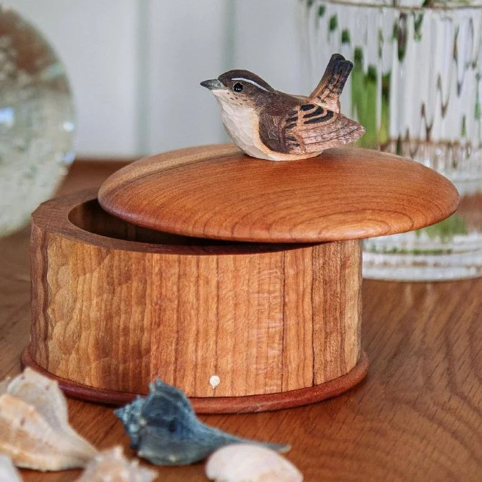 Ronde houten bewaardoos met een bruin winterkoninkje als dekselgreep, gefotografeerd op een houten tafel met decoratieve schelpen.
