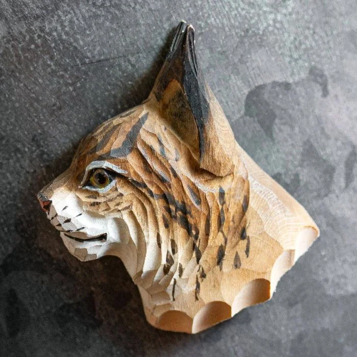 Houten lynxkop als magneet op een donkere metalen wand, met lange zwarte oorpunten, bruingevlekte kop en lichte snuit met donkere stippen.