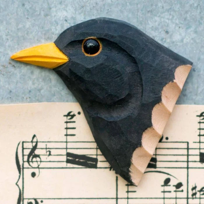 Decoratieve houten vogelmagneet in de vorm van een merel, met helder gele snavel en zwarte veren, liggend op een muziekblad met noten.