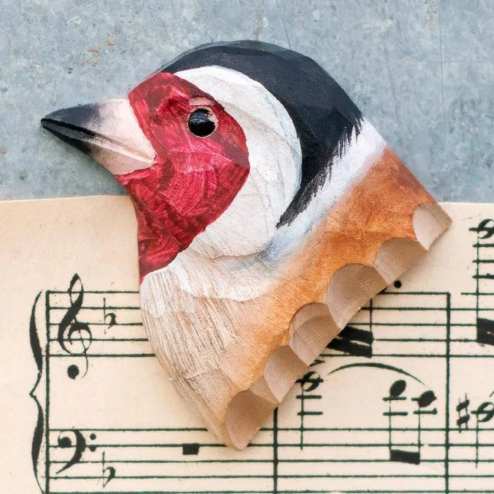 Decoratieve houten vogelmagneet in de vorm van een putter, met opvallend rood gezicht en zwart-witte kop, liggend op een muziekblad.