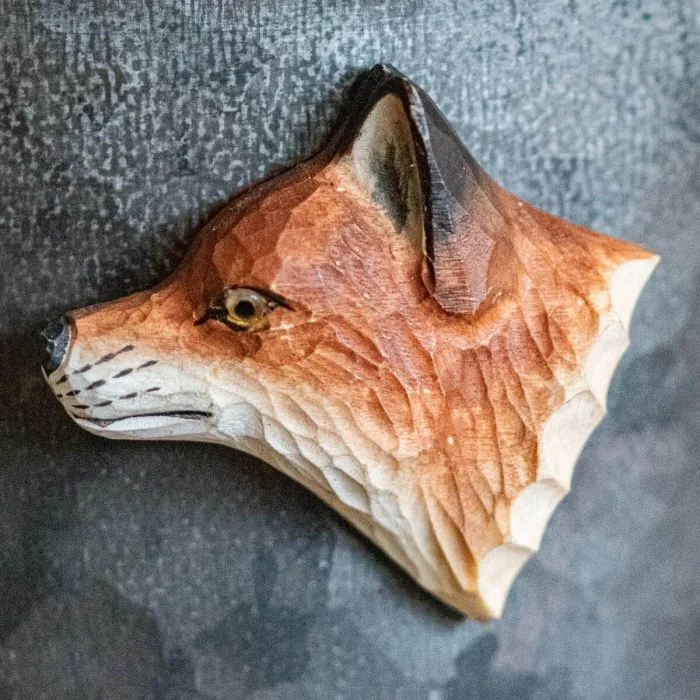 Beschilderde houten vossenkop als magneet op een grijze metalen wand, met amberkleurig oog, donkere oorrand en lichtgekleurde vacht langs de hals.