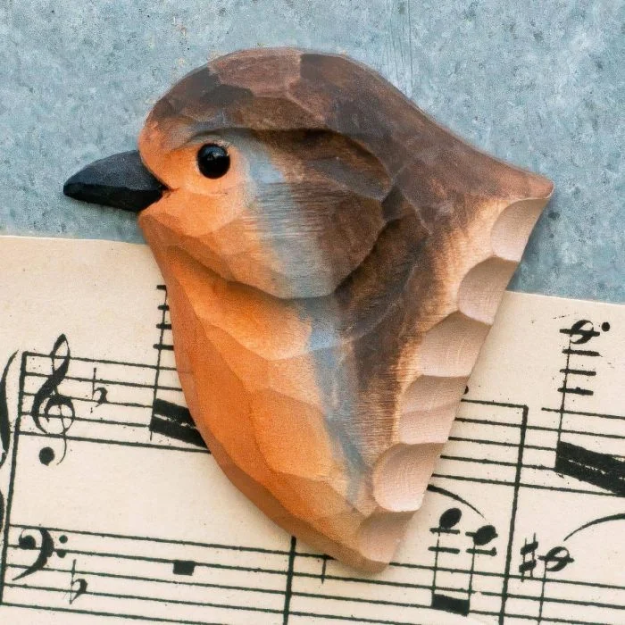 Decoratieve houten vogelmagneet in de vorm van een roodborstje, met opvallende oranje borst en donkere snavel, liggend op bladmuziek met muzieknoten.