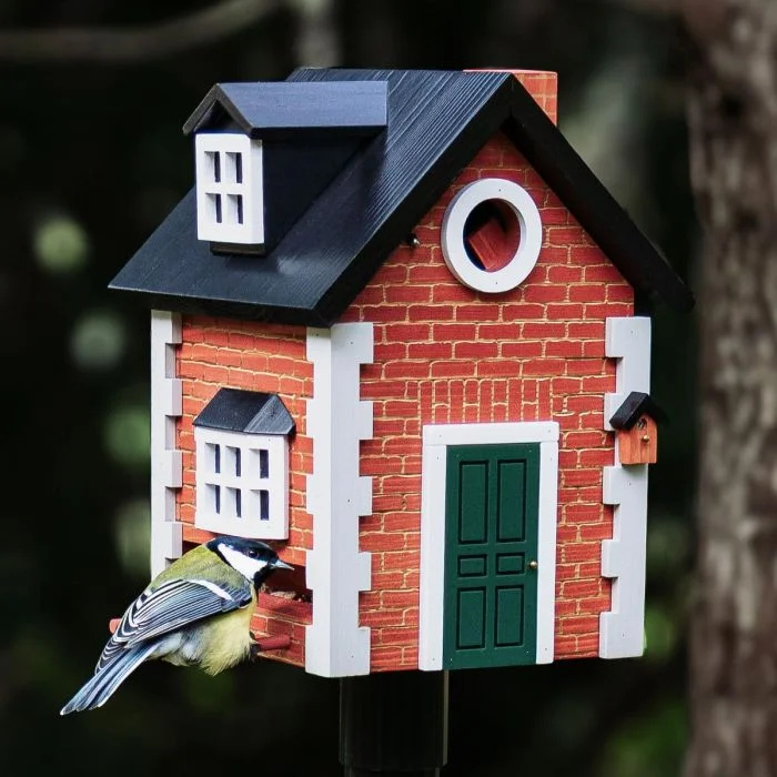 Houten vogelhuis in Engelse cottage-stijl met een koolmees op de zitstok, rode baksteenprint, donkergroene deur en witte ramen tegen een donkere tuinachtergrond.